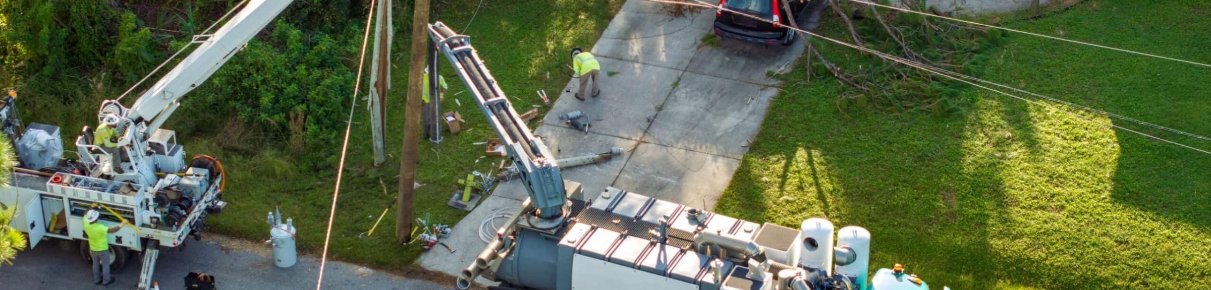 Utility crews with bucket and digger-derrick trucks resetting a pole beside a storm-downed tree, seen from above